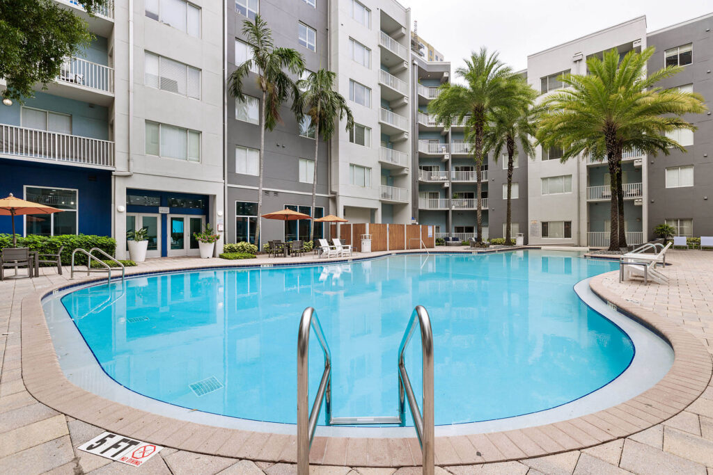 Apartment complex pool surrounded by lounge chairs, umbrellas, palm trees, and multi-story gray buildings with balconies and windows. The pool has metal railings and a 5 FT depth marker at the entrance.