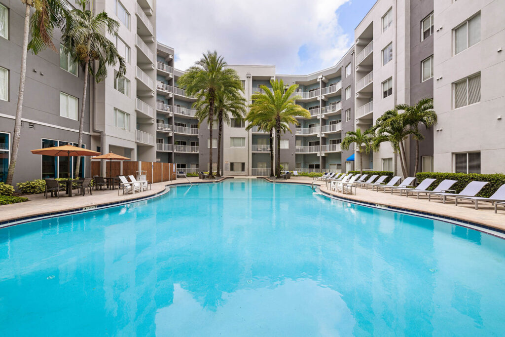 A modern apartment complex with a large, clear swimming pool surrounded by lounge chairs, palm trees, and shaded seating areas on a sunny day.