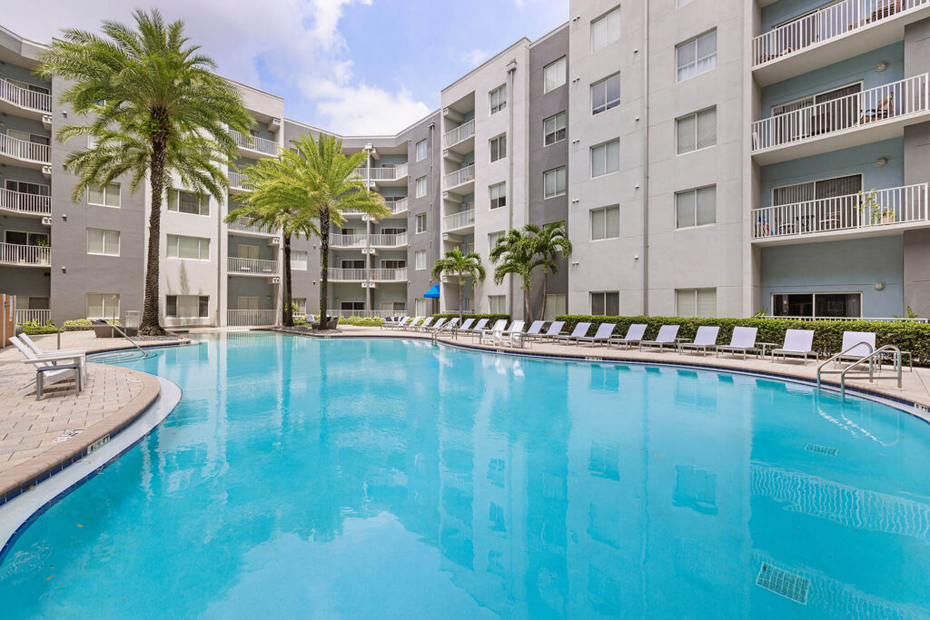 A large outdoor swimming pool surrounded by lounge chairs, palm trees, and a multi-story light gray apartment building under a partly cloudy sky.