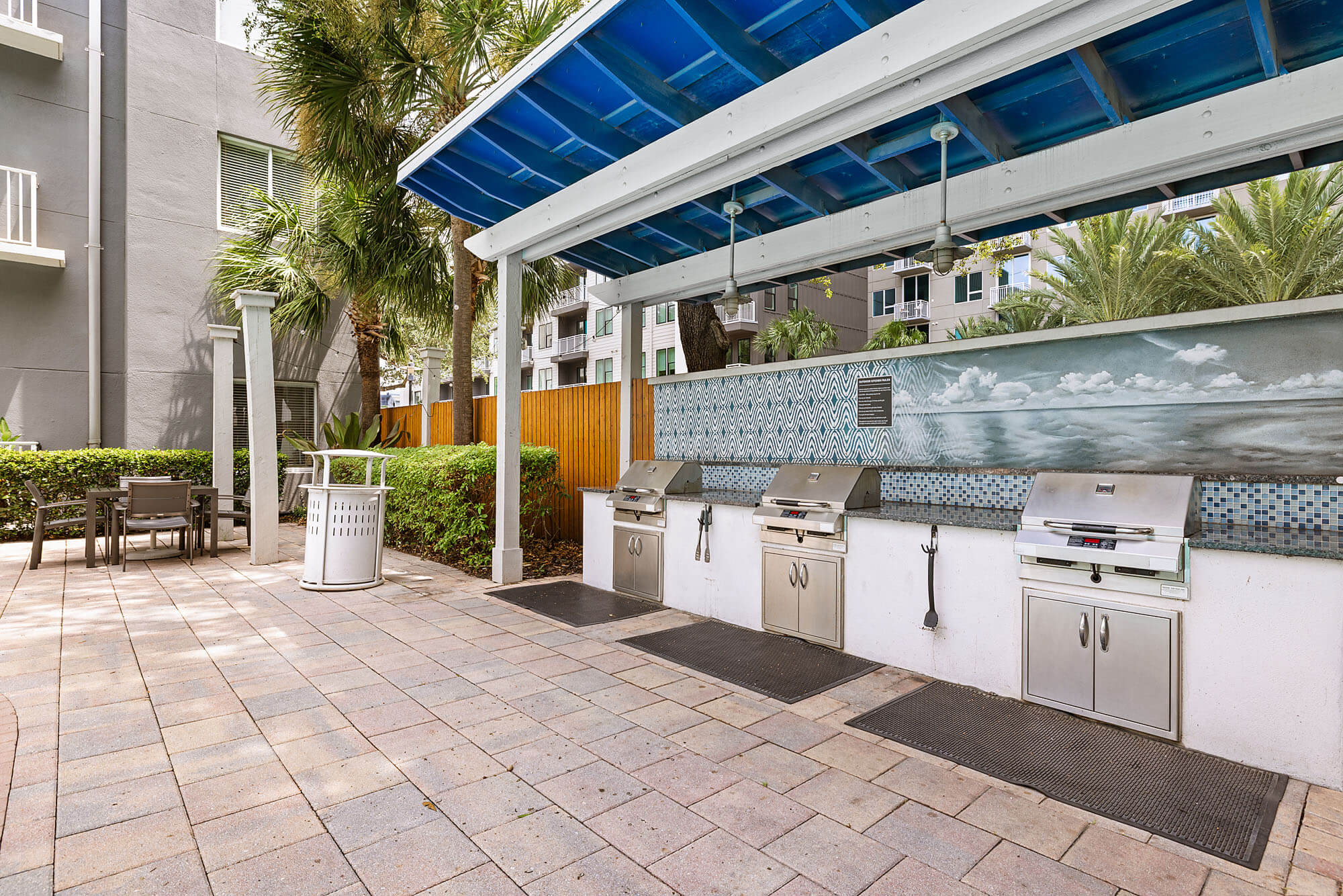 Outdoor grilling area with three stainless steel grills under a blue-roofed canopy, nearby trash can, table, and chairs; modern apartment buildings and palm trees in the background.