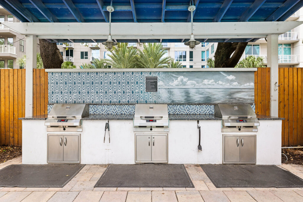 Three stainless steel outdoor grills under a blue canopy, with a patterned tile backsplash and a beach mural. Residential buildings and palm trees are visible in the background.