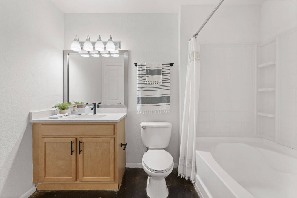 Bright bathroom with a wooden vanity, white countertop, round toilet, striped towel on a rack, and a white shower-tub combo with a shower curtain, under four-bulb vanity lighting.