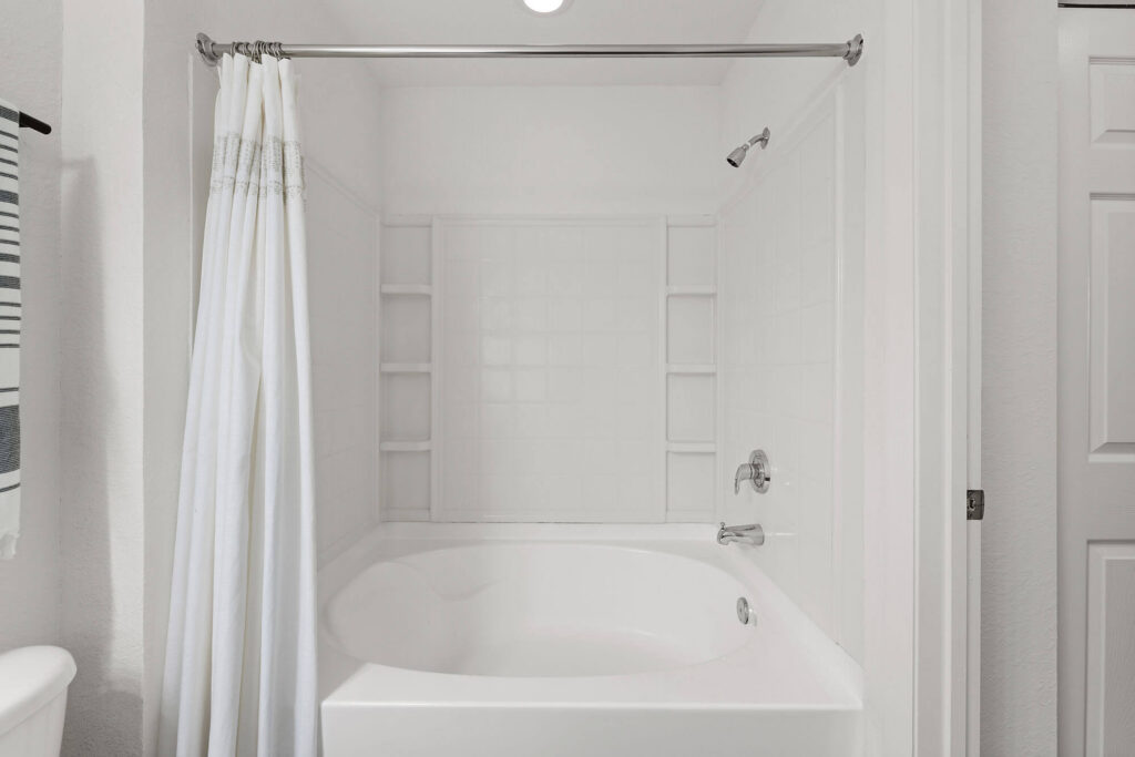 White bathroom with a bathtub and shower, white tile walls, built-in shelves, a curved shower curtain rod with a white curtain, and a towel hanging on the left. The space appears clean and minimalistic.