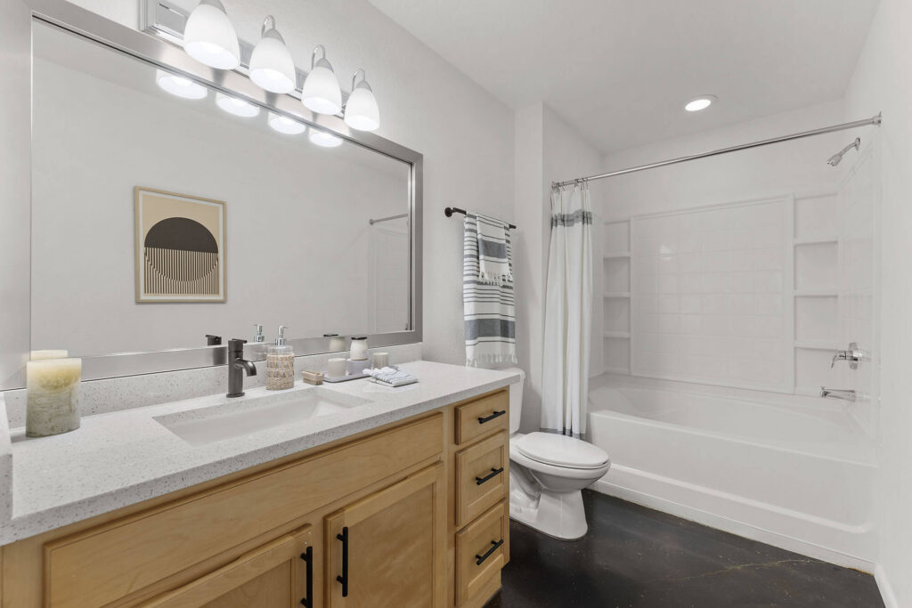 Modern bathroom with light wood vanity, double sink, large mirror with four lights, white countertop, toilet, striped shower curtain, white bathtub, and shelf wall art. Towels, soap, and candles are on the counter.