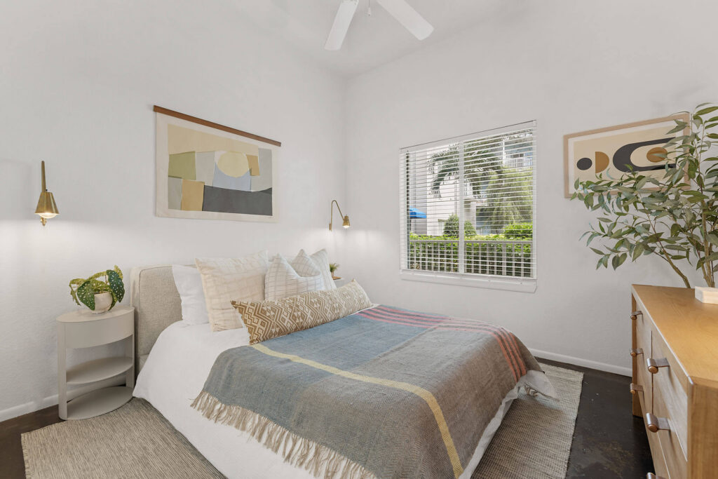 Bright bedroom with a bed covered by a patterned blanket, pillows, wall art, bedside lamps, a small plant, a window with blinds, and a dresser with a potted plant.