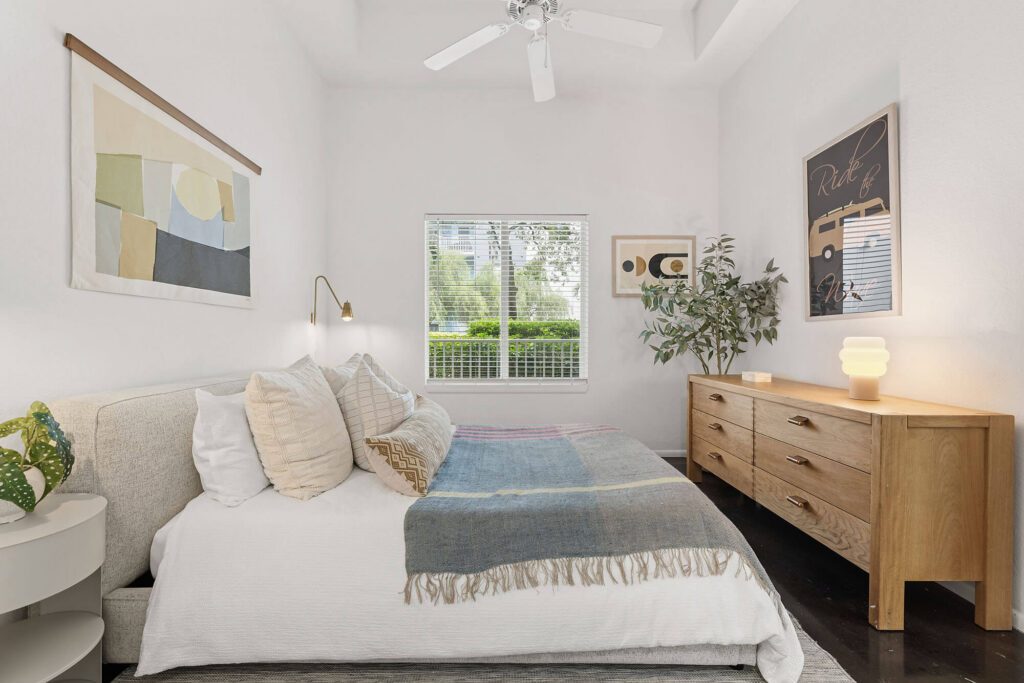Bright bedroom with a bed, cushions, and a striped blanket, wooden dresser, potted plants, wall art, and a window letting in natural light; white walls and ceiling fan above.