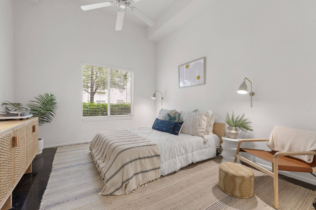 A cozy bedroom with a bed, striped bedding, a wooden chair, nightstands, wall lamps, a large window, indoor plants, and neutral decor.