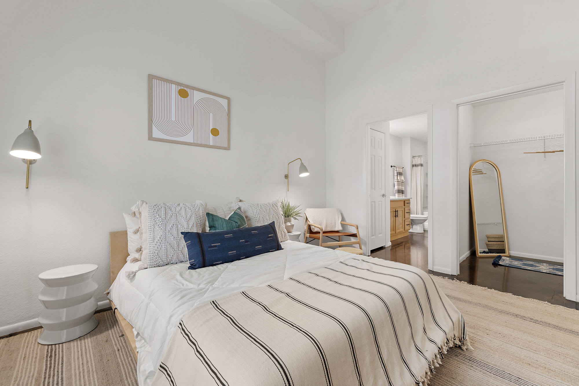 A cozy bedroom with a bed covered in white and striped bedding, pillows, wall lamps, a side table, a chair, modern art above the bed, and an open door to a walk-in closet with a mirror and a view into a bathroom.