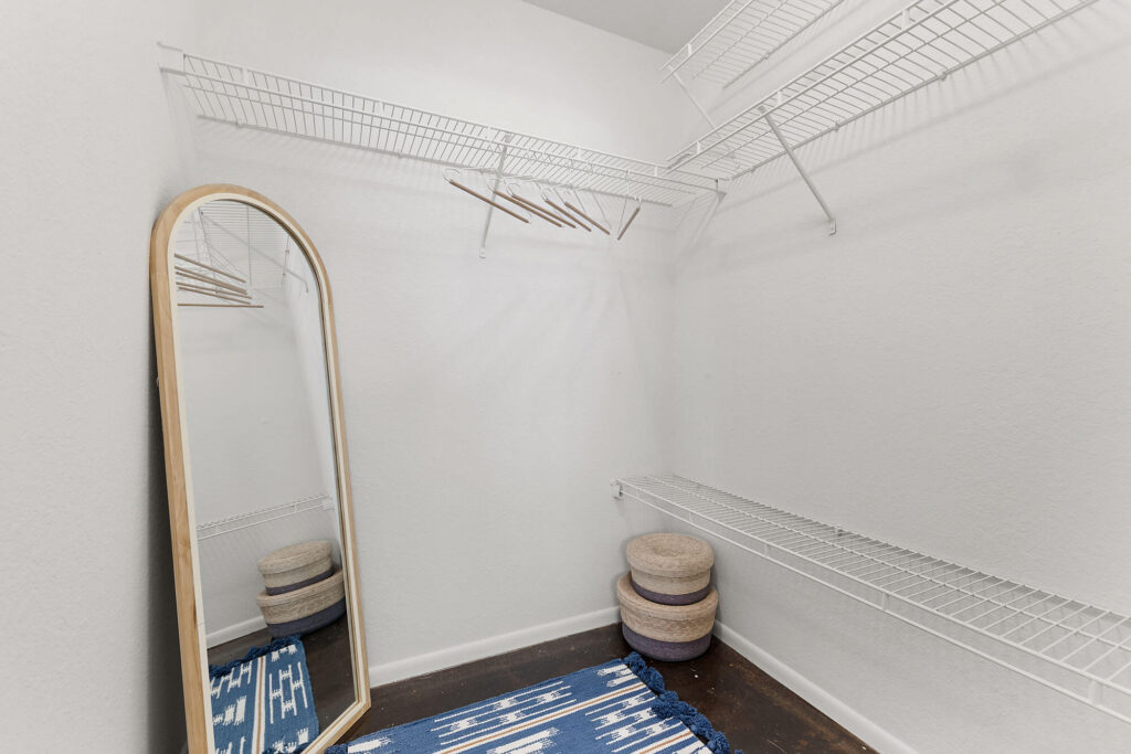 An empty walk-in closet with white wire shelves, wooden hangers, a tall oval mirror, stacked round baskets, and a blue patterned rug on a dark floor.
