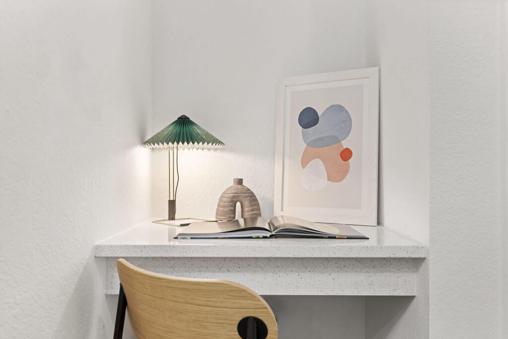 A small desk with a wooden chair, green desk lamp, ceramic vase, open book, and abstract art in a white frame on a white surface against white walls.