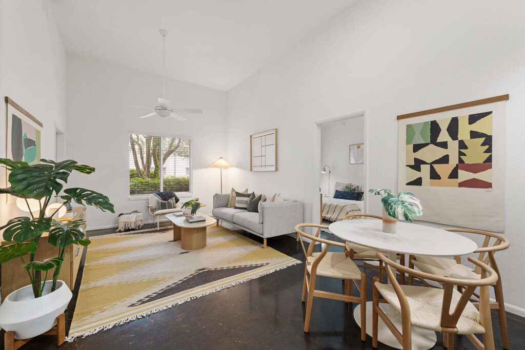 Bright living room with a beige sofa, round dining table, abstract art, indoor plants, large window, and a yellow patterned rug on a dark floor. Ceiling fan and soft lamp add light.