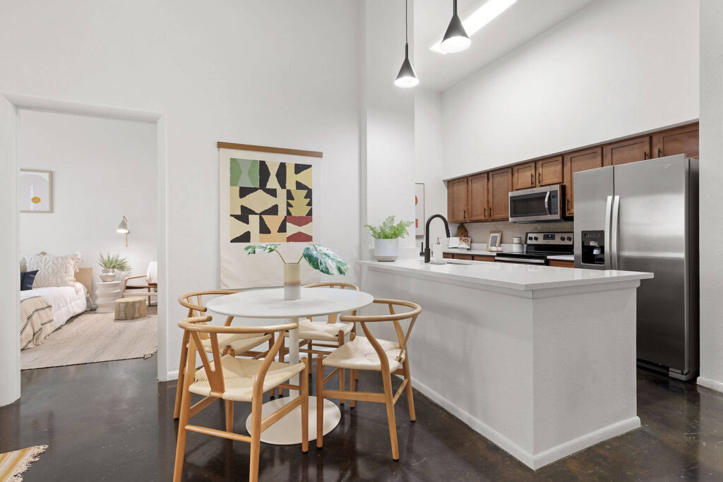 Modern kitchen and dining area with wooden chairs, round table, stainless steel appliances, white countertops, abstract wall art, and view into a cozy bedroom with neutral decor.
