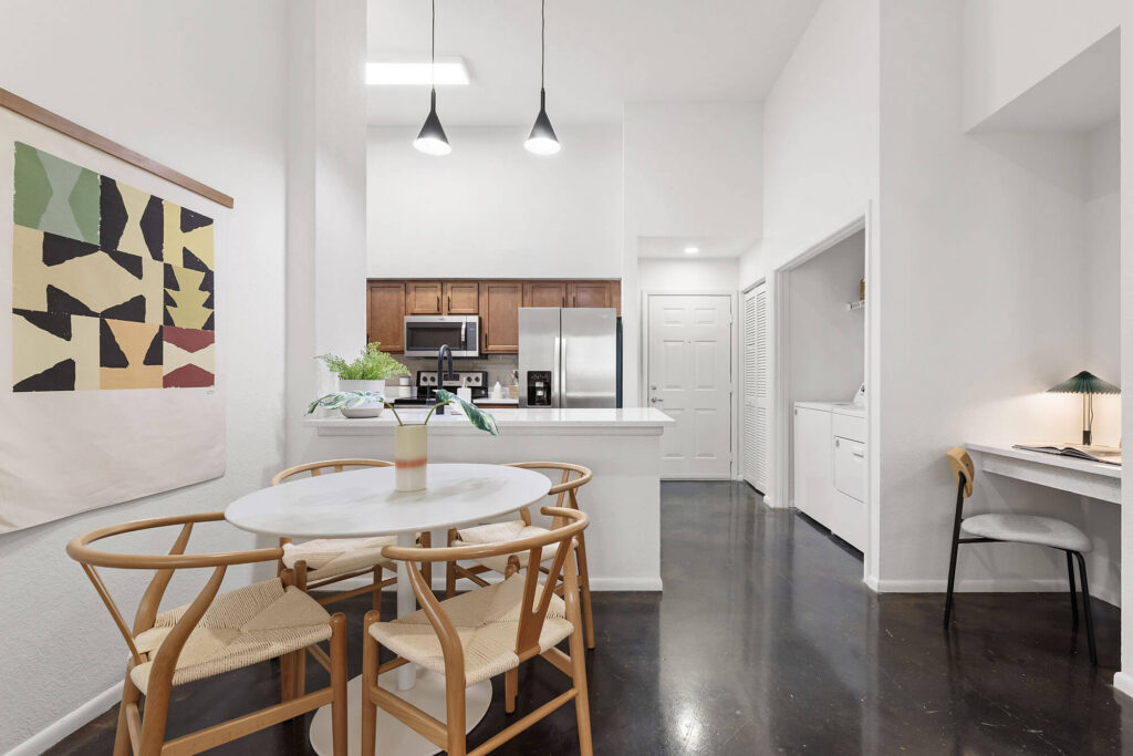 Modern kitchen and dining area with wood cabinets, round white table with four chairs, minimalist art on the wall, laundry area, and small desk with a lamp, all in a bright, white interior.