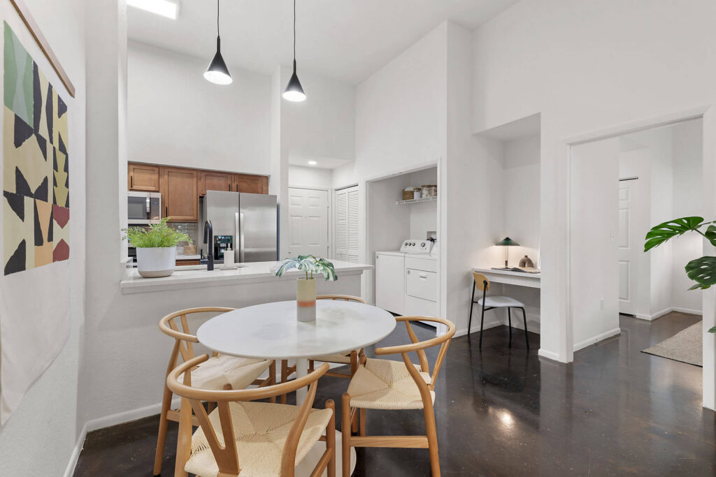 A modern dining area with a round table and four chairs, next to an open kitchen with stainless steel appliances. A small workspace and laundry nook are visible, with plants and wall art as decor.