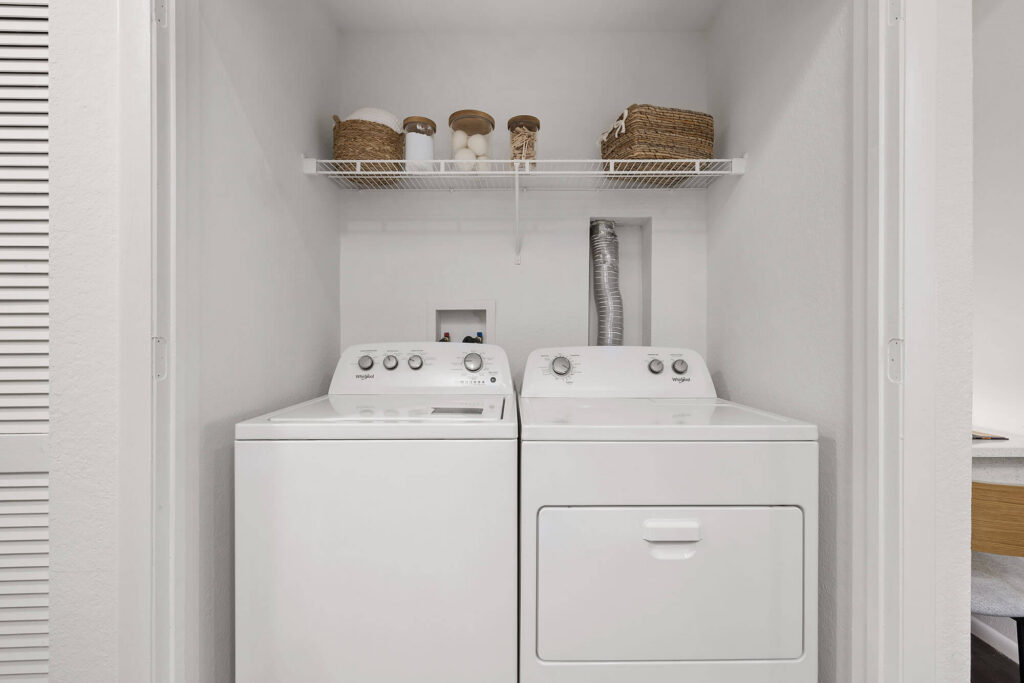 A laundry closet with a white washer and dryer below a wire shelf holding baskets, jars, and bottles, all set against white walls.