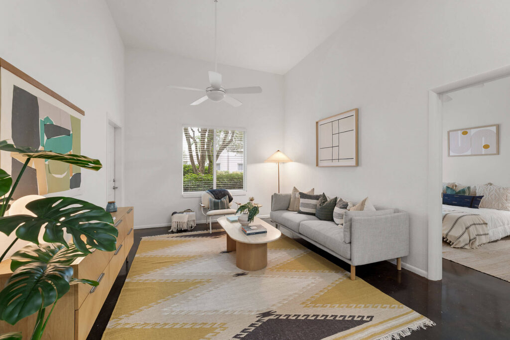 Modern living room with a gray sofa, abstract art, large indoor plant, and geometric rug. Window with outdoor view, ceiling fan, and adjacent bedroom visible through open doorway. Neutral tones and minimalist decor.