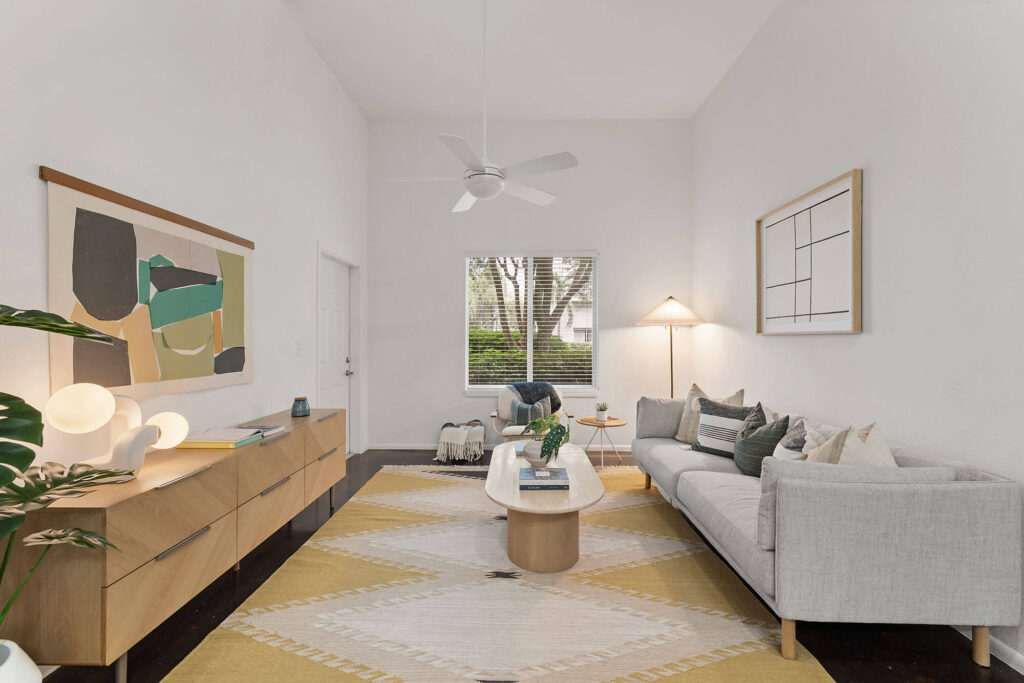 Modern living room with a gray sofa, yellow patterned rug, wooden coffee table, sideboard, abstract art, floor lamp, ceiling fan, and window with a view of trees outside.