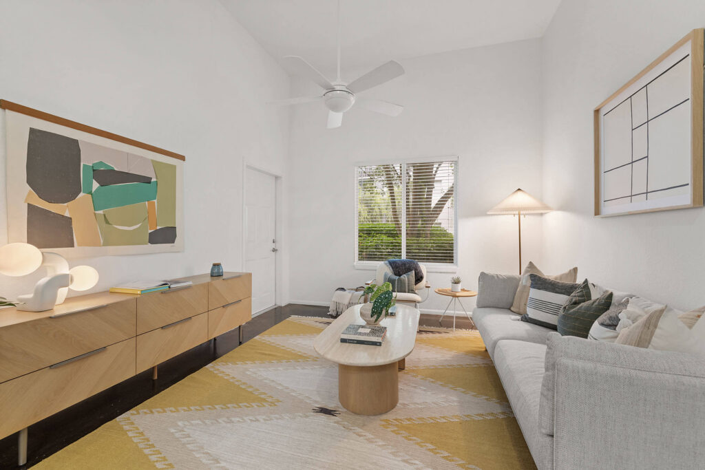 Modern living room with a gray sofa, wooden coffee and side tables, abstract wall art, a large window with blinds, and a yellow patterned rug on a dark floor. A floor lamp and ceiling fan are also visible.