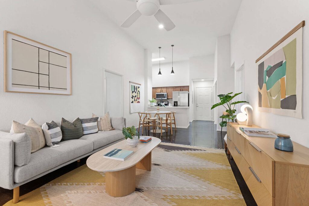 Modern living room with a light gray sofa, wooden coffee table, and geometric rug. Open kitchen and dining area in the background, ceiling fan overhead, and abstract art on white walls.