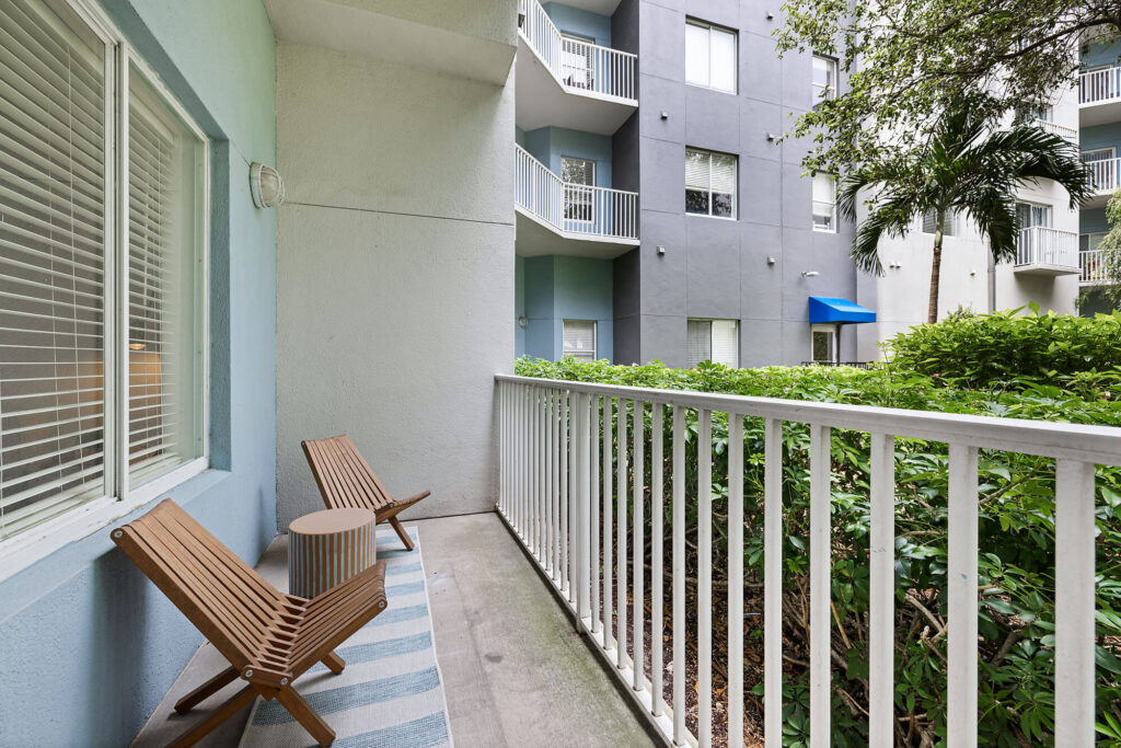 A small apartment balcony with two wooden chairs, a side table, and a striped rug, overlooking green bushes and nearby apartments with white railings.