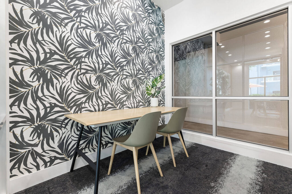 A modern office space with a light wood table, two gray chairs, black and white leaf-patterned wallpaper, a plant, and large interior windows.