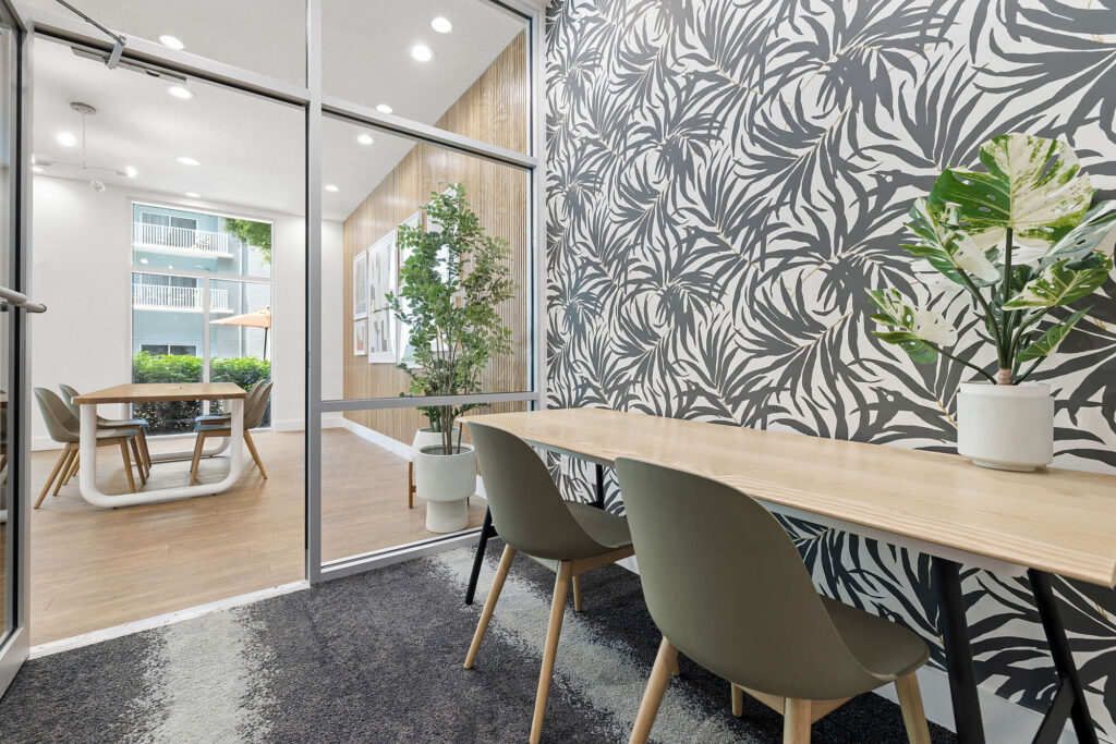 Modern office space with glass walls, two green chairs at a wooden table, and a bold black-and-white leaf-patterned accent wall. Potted plants add greenery to both rooms. Natural light brightens the area.