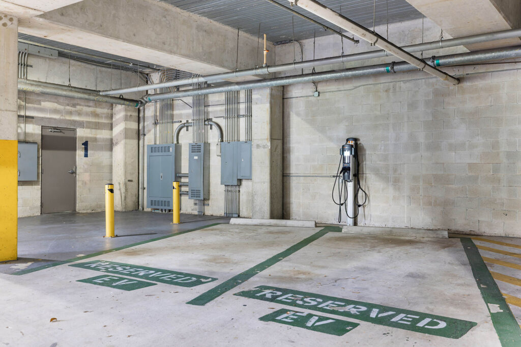 Two parking spaces in a concrete parking garage are marked Reserved EV. An electric vehicle charging station is mounted on the wall between the spaces. Yellow poles separate the area from the rest of the garage.