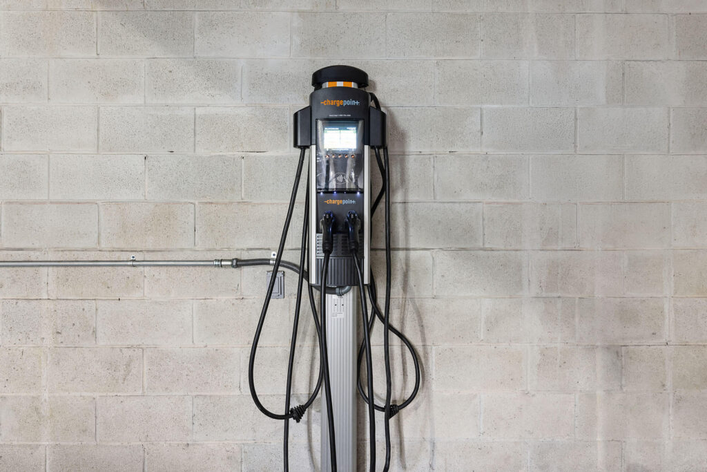 An electric vehicle charging station is mounted on a concrete block wall, with cables and connectors hanging down from the unit.