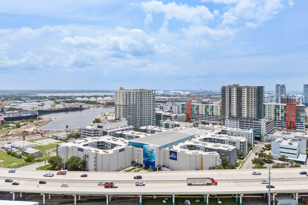 Aerial view of modern apartment buildings, a highway with vehicles, a river, and an industrial area in the background under a partly cloudy sky.