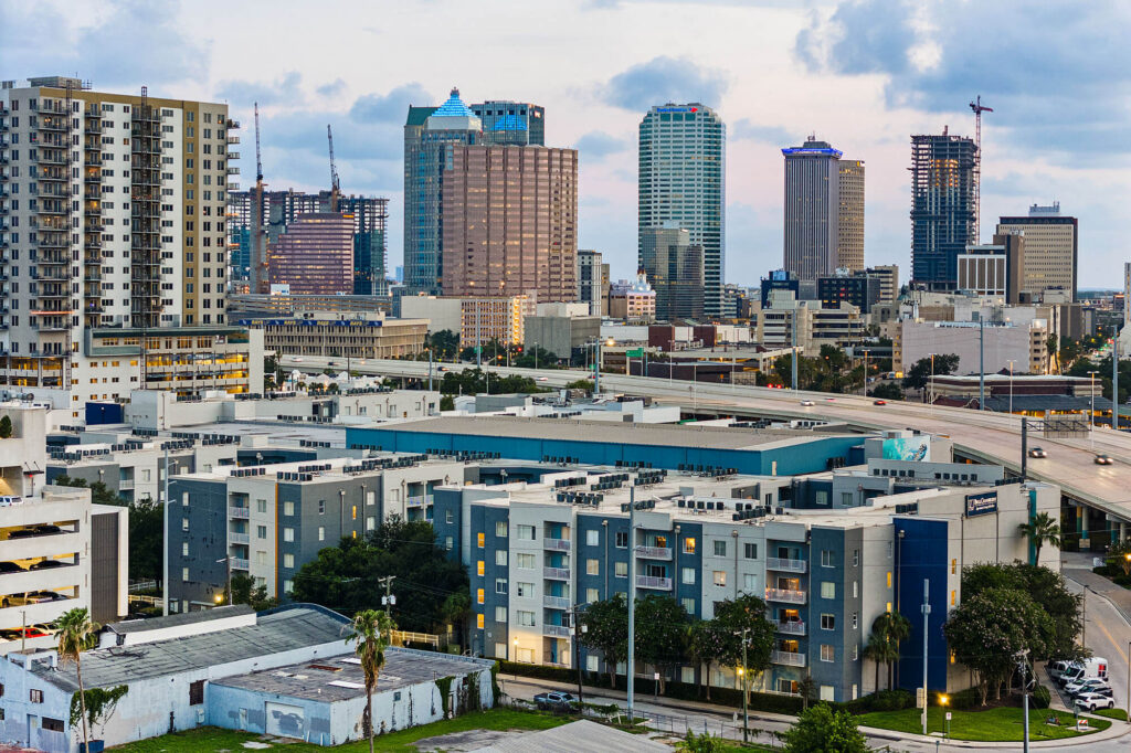 A cityscape with modern high-rise buildings in the background and mid-rise apartment complexes with blue accents in the foreground, under a cloudy sky during daylight.