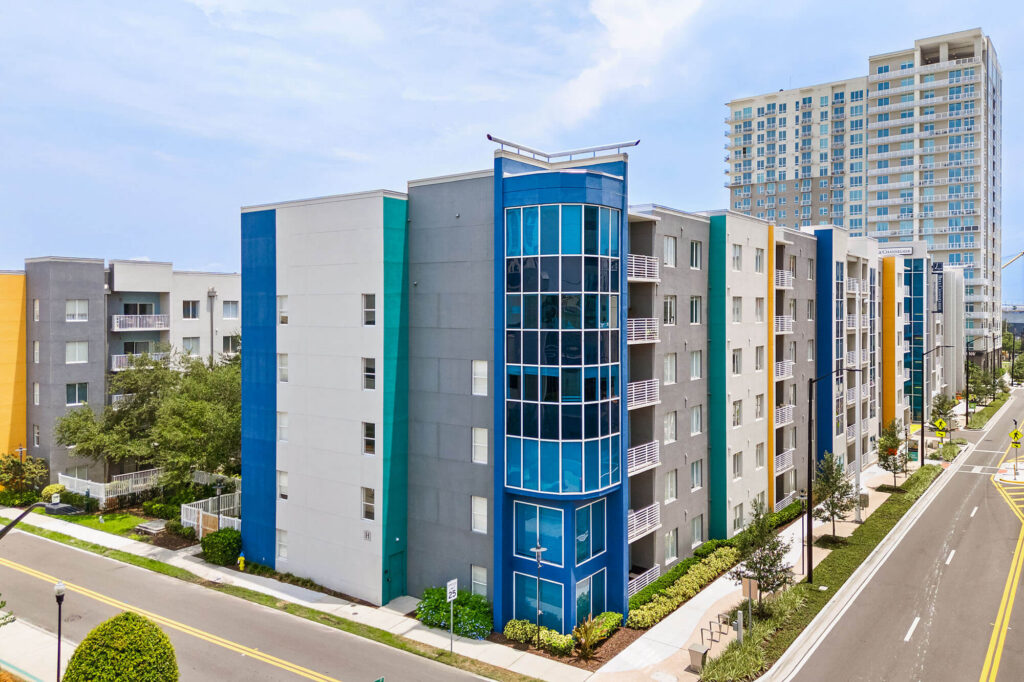 Modern mid-rise apartment buildings with colorful facades line a clean city street, with trees and a sidewalk in front. A taller high-rise is visible in the background under a partly cloudy sky.
