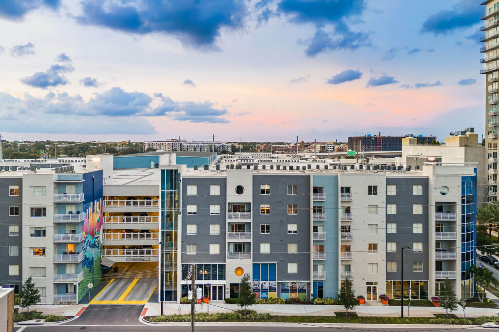 A modern mid-rise apartment building with colorful mural, balconies, and large windows is shown at sunset. A taller building stands to the right, with cityscape and cloudy sky in the background.