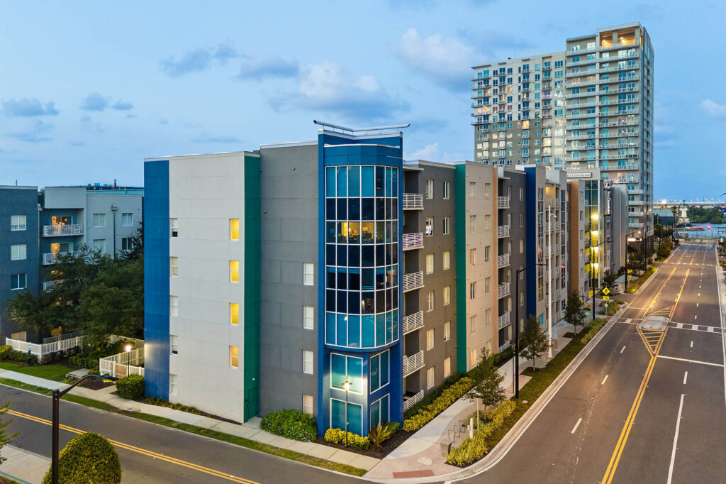 Modern mid-rise apartment buildings line a quiet, empty street at dusk, with a tall high-rise in the background and soft street lighting illuminating the scene.