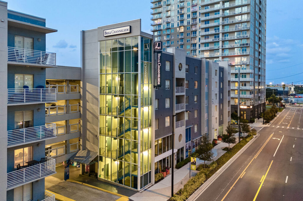 Modern apartment buildings line a quiet city street at dusk, with illuminated glass stairwells and balconies visible. Streetlights are on, and a tall high-rise building stands in the background.