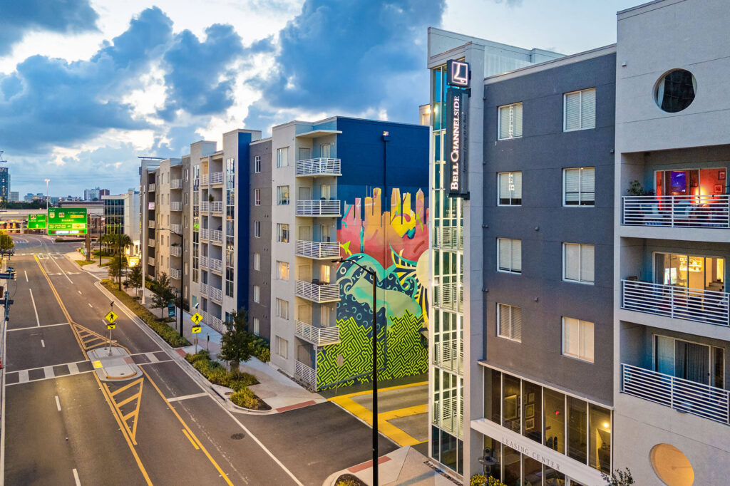 Modern apartment buildings line a city street at sunset; a vibrant mural with colorful hands and patterns decorates one buildings wall. Balconies, trees, and street signs are visible, with cars absent from the road.