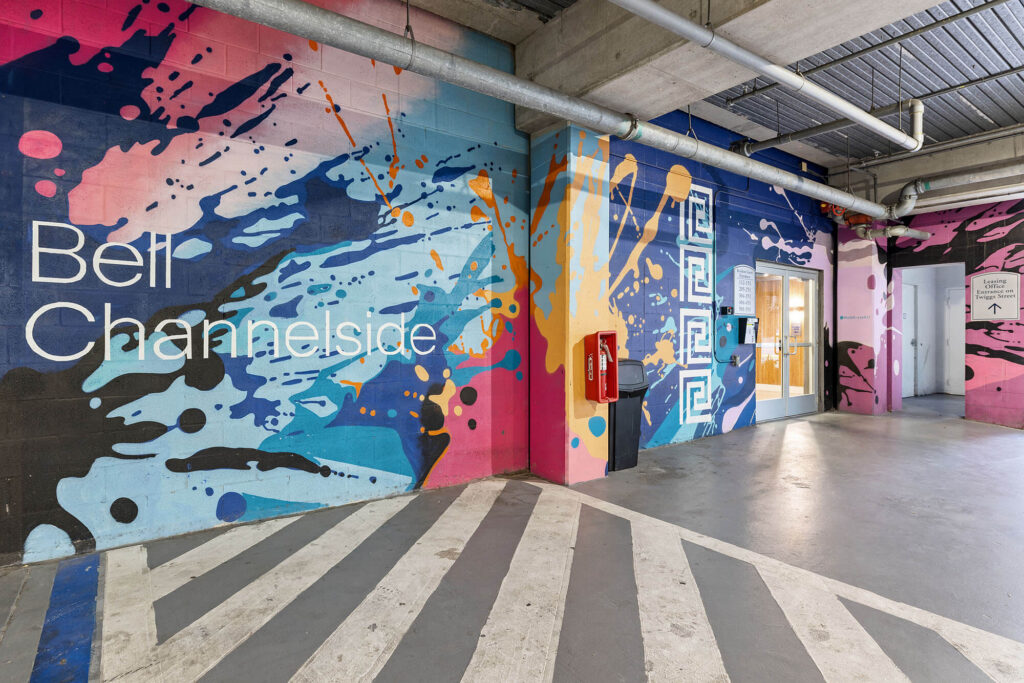 Colorful mural with abstract splashes and Bell Channelside text on a parking garage wall; fire extinguisher, door, and elevator are visible nearby.