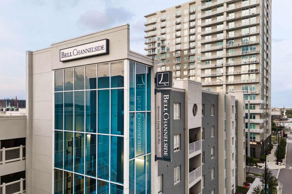Modern apartment building with large blue-tinted windows and Bell Channelside signs on the exterior. A taller residential tower stands nearby, and a city street runs along the buildings.