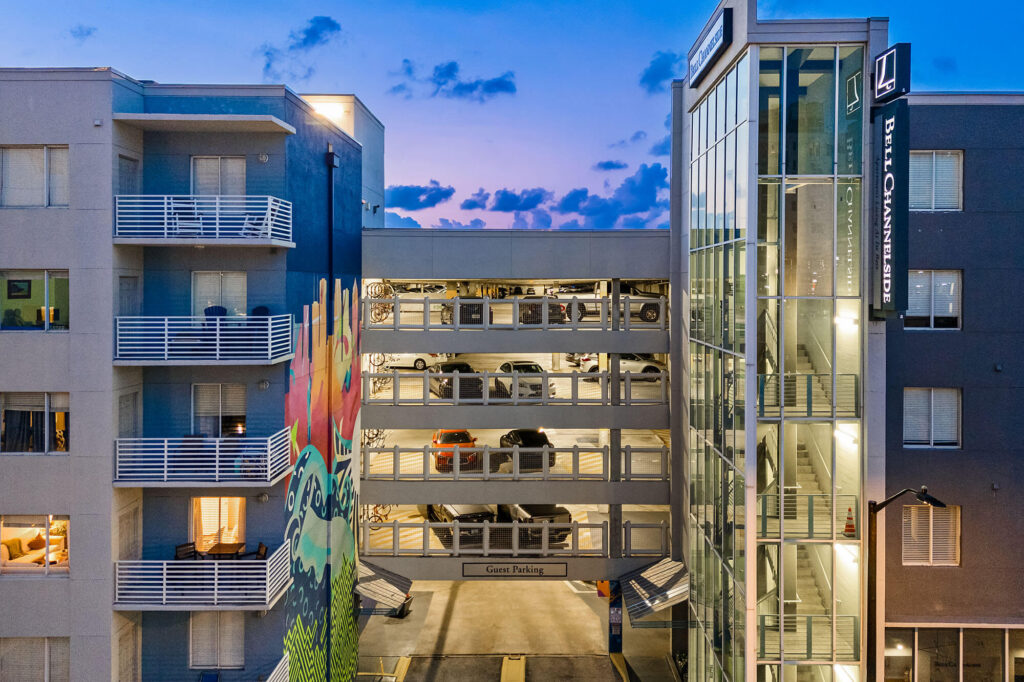Modern apartment complex at dusk with a colorful mural, glass elevator shaft, and a multi-level parking garage. Some lights are on in the building, and a few cars are parked inside the garage.
