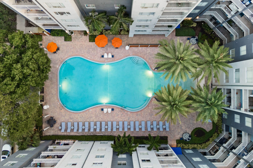 Aerial view of a kidney-shaped swimming pool surrounded by lounge chairs, palm trees, and orange umbrellas, set within a modern apartment complex.