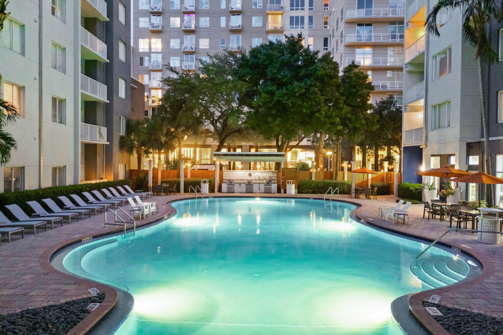 A large illuminated outdoor pool surrounded by lounge chairs, trees, and apartment buildings at dusk, with a bar area in the background and umbrellas to the right.