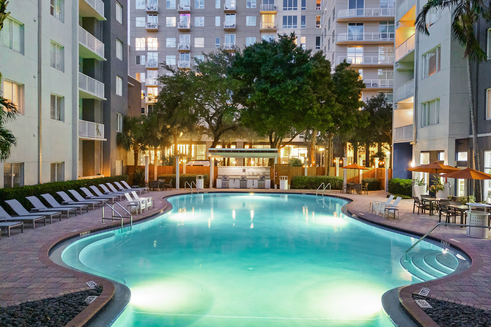 A large illuminated outdoor pool surrounded by lounge chairs, trees, and apartment buildings at dusk, with a bar area in the background and umbrellas to the right.