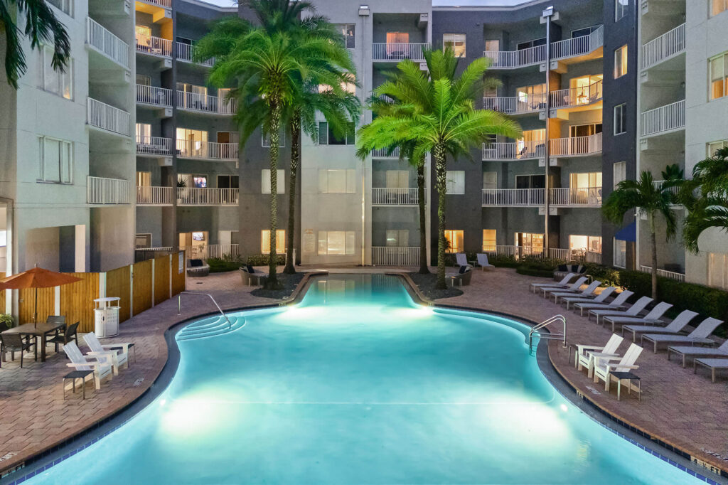 A brightly lit swimming pool surrounded by lounge chairs and palm trees sits in the courtyard of a modern apartment complex in the evening.